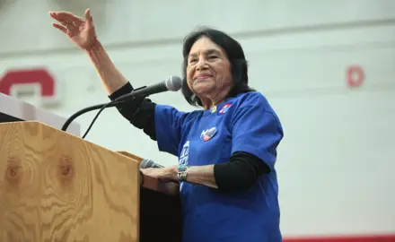 Dolores Huerta stands at a podium. She is smiling, and her right arm is lifted toward the crowd.