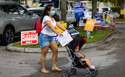 A Latina woman wearing a mask pushes her child in a stroller, with several political campaign signs behind her in the background.