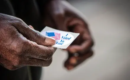 A close up of an elderly Black man's hands. He has just finished casting his ballot, and is holding his “I voted” sticker.