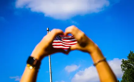 Two hands form a hard with their fingers in front of an American flag and blue sky.