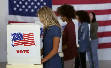 Four women cast ballots at voting booths with an American flag in the background.