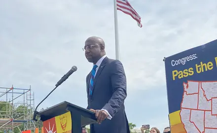 Sen. Raphael Warnock standing at a podium outdoors with an American flag in the background.