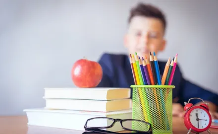 A child sitting at a desk with books, an apple, glasses, a clock, and rainbow pencils in a cup on the desk.