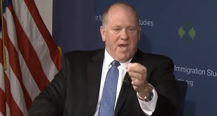 Tom Homan, in suit and tie and seated in front of an American flag, gestures while speaking. 