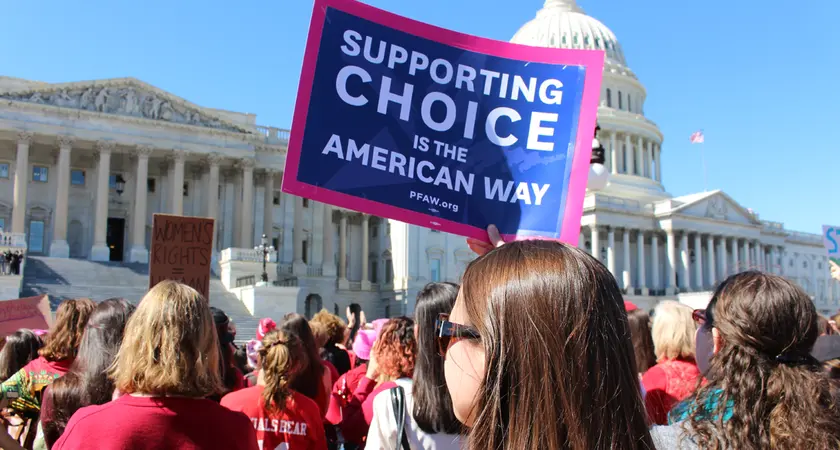 A group of people stand in front of the Capitol Building; one woman holds a sign that says "Supporting Choice is the American Way"