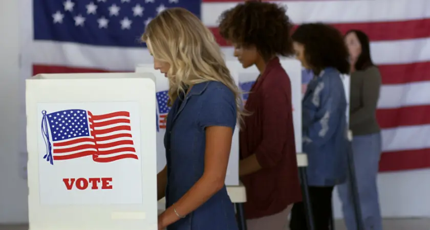 Four women cast ballots at voting booths with an American flag in the background.