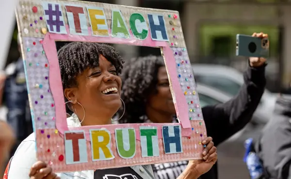 Woman holding a sign framing her face  that says #TeachTruth