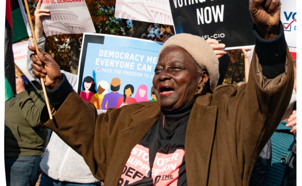 Older woman at a protest in front of a sign that says "voting rights now"