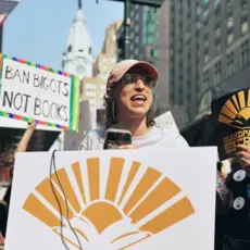 Alana Byrd speaking at a rally for Grandparents For Truth with people holding signs in the background.