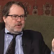 John Zmirak, a rumpled white guy sitting in an armchair, gestures with his hand while speaking with Eric Metaxas, who is off-camera. 