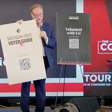 Richard Rogers stands on stage holding a placard with a QR code for voter registration. Logo of the Courage Tour is visible behind him. 