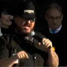 Stewart Rhodes, wearing a cowboy hat emblazoned with Oath Keepers logo," speaks into microphone held in his left hand while gesturing with his right. Rally emcee Eric Metaxas is visible behind him. 