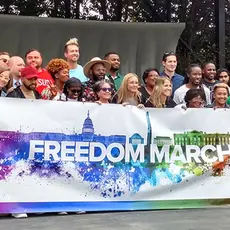 A couple dozen speakers and singers gather on stage behind a rainbow-colored "Freedom March" banner.