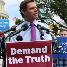 Man in tie speaking at podium in front of crowd. Podium has sign that says Demand the Truth.