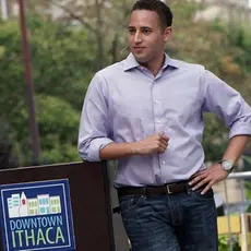 Svante Myrick wearing a purple shirt and standing next to a podium which says "Downtown Ithaca."