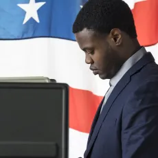 A Black man in a suit stands at a voting booth with an American flag in the background.