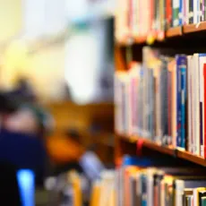 A full bookshelf in the foreground and people in a library in the background.