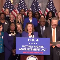 Nancy Pelosi stands at a podium with a group of people behind her while she talks about the Voting Rights Advancement Act