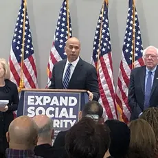 A group of lawmakers including Cory Booker and Bernie Sanders stand behind a podium with a sign that says "Expand Social Security"