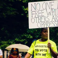 A person wearing a shirt that says "Register to Vote with Me" carries a sign that says "No one is free while others are oppressed."