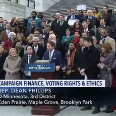 A screenshot from C-SPAN2 where a group of people stand behind Rep. Dean Phillips on a set of stairs as he talks about campaign finance, voting rights, and ethics.