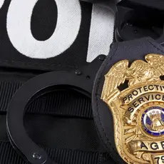 Closeup of a metal badge that says "Protective Services Agent" on a police vest.