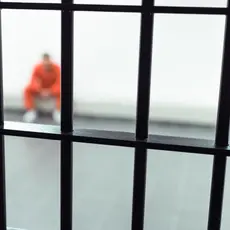 An image of a prisoner in an orange jumpsuit shown seated in a corner behind iron bars