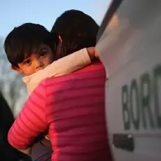 A young child clings to his mother near a border patrol truck and agent.