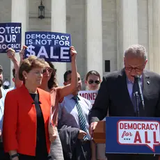 Chuck Schumer speaks at a podium while people around him hold signs saying "Protect our democracy" and "Democracy is not for sale."