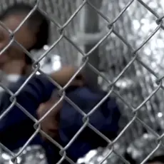 Children wrapped in emergency blankets laying behind a chain-link fence.