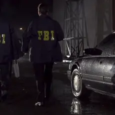 Two men in FBI jackets walk past a black car at night.