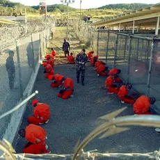 Detainees in orange jumpsuits sit on their knees and face a chain link fence at Camp X-Ray in Guantanamo Bay