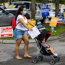 A Latina woman wearing a mask pushes her child in a stroller, with several political campaign signs behind her in the background.