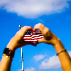 Two hands form a hard with their fingers in front of an American flag and blue sky.