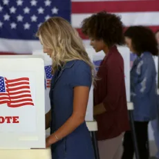 Four women cast ballots at voting booths with an American flag in the background.