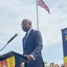 Sen. Raphael Warnock standing at a podium outdoors with an American flag in the background.