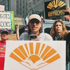 People at a rally. Older woman holds a black sign with yellow letters that reads "grandparents for truth"