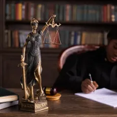 A Black female judge writes at a desk with a statue of Lady Justice on it.