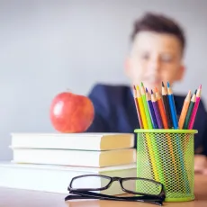 A child sitting at a desk with books, an apple, glasses, a clock, and rainbow pencils in a cup on the desk.